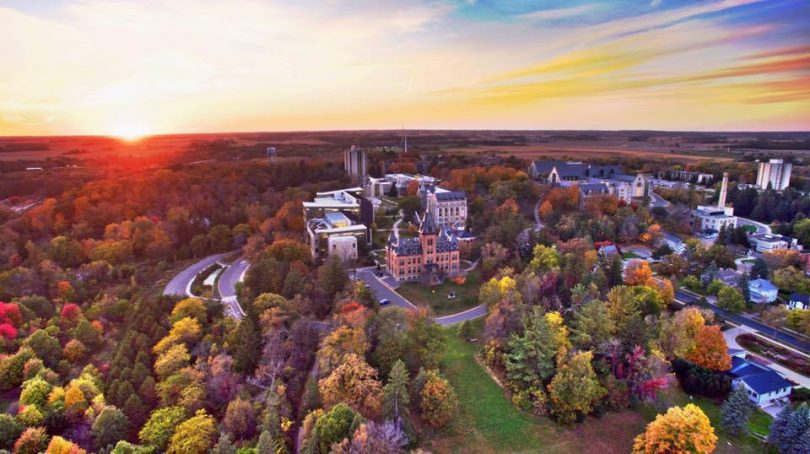 St Olaf College seen from an aerial view.  The campus is surrounded by deciduous trees in early fall bloom, and the sun is shining.
